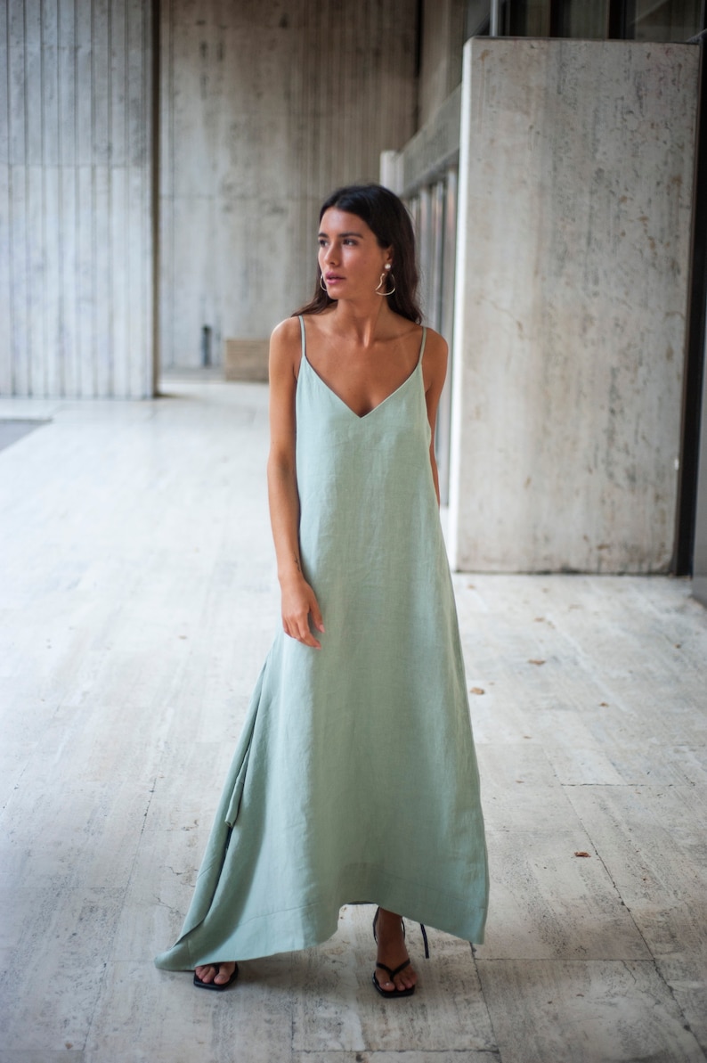 May include: A woman wearing a light green maxi dress with a V-neckline and thin straps. The dress has a flowing, asymmetrical hemline. She is standing in a concrete hallway.