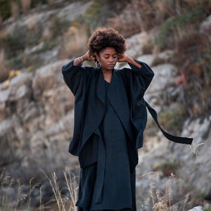May include: A woman wearing a black long-sleeved dress with a flowing skirt and white boots. She is standing on a rocky surface with green plants in the background.