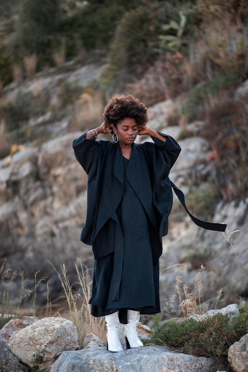 May include: A woman wearing a black long-sleeved dress with a flowing skirt and white boots. She is standing on a rocky surface with green plants in the background.