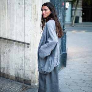 May include: A woman wearing a grey and white patterned jacket with a long grey skirt. She is standing in front of a white wall and looking over her shoulder.