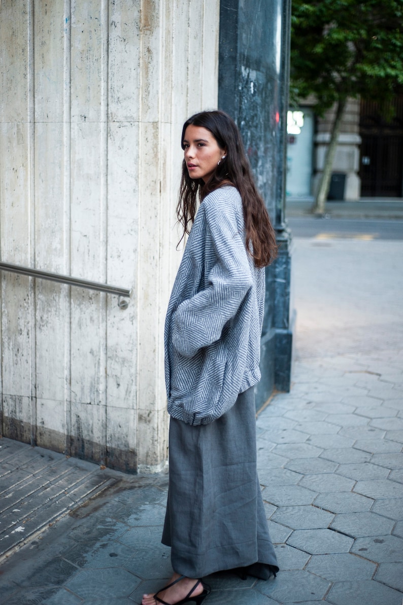 May include: A woman wearing a grey and white patterned jacket with a long grey skirt. She is standing in front of a white wall and looking over her shoulder.