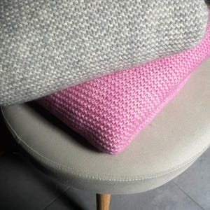 May include: Two knitted throw pillows, one gray and one pink, are stacked on a light beige stool with wooden legs.