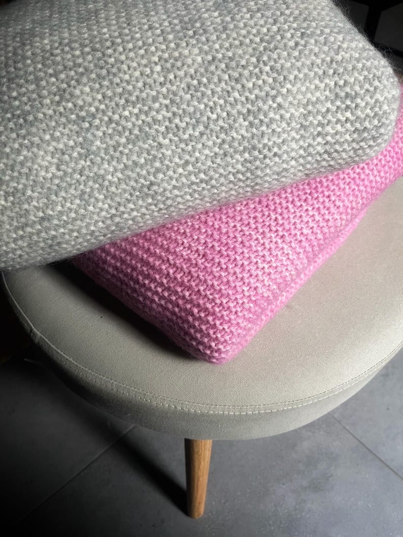 May include: Two knitted throw pillows, one gray and one pink, are stacked on a light beige stool with wooden legs.