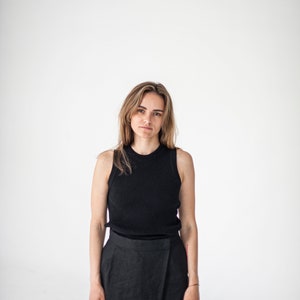 Op de afbeelding: Een vrouw draagt een zwart tanktop en een zwarte wikkelrok. Ze staat voor een witte achtergrond.