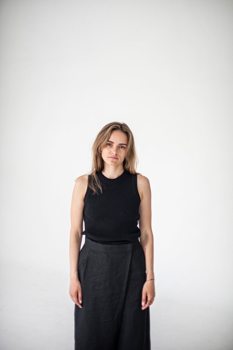 Op de afbeelding: Een vrouw draagt een zwart tanktop en een zwarte wikkelrok. Ze staat voor een witte achtergrond.