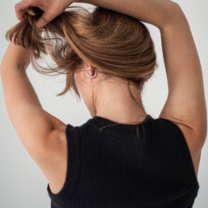 Op de afbeelding: Een vrouw met lang bruin haar draagt een zwart topje zonder mouwen. Ze houdt haar haar omhoog met haar rechterhand.