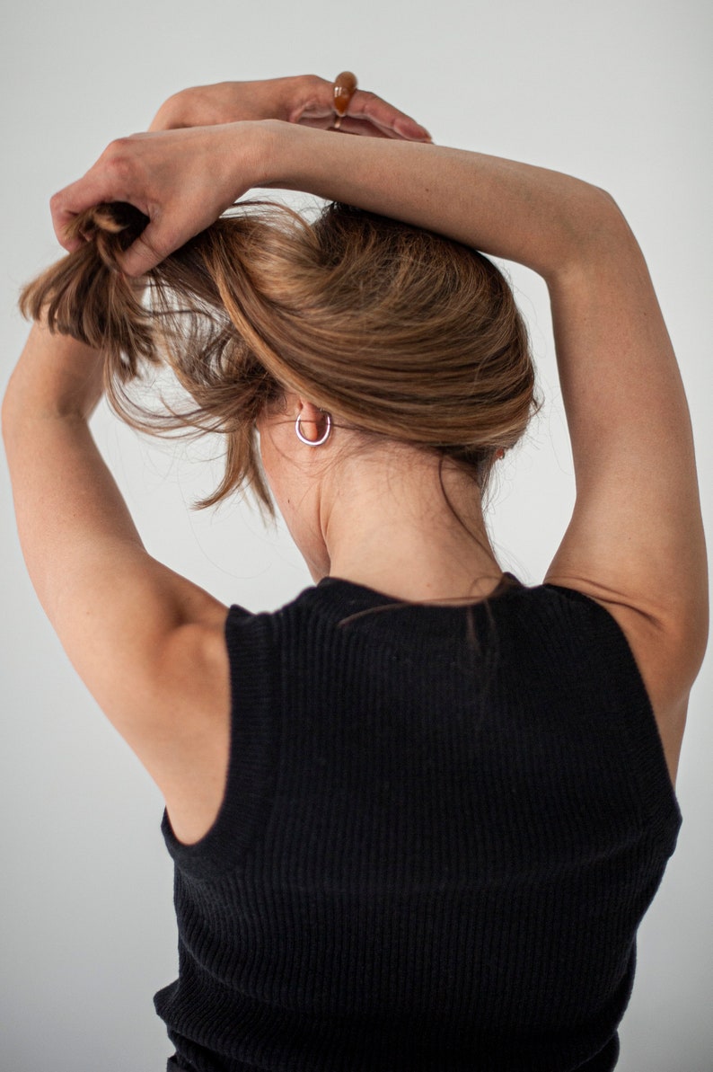 Op de afbeelding: Een vrouw met lang bruin haar draagt een zwart topje zonder mouwen. Ze houdt haar haar omhoog met haar rechterhand.