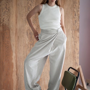 Op de afbeelding: Een vrouw draagt een wit geribbeld tanktop en een lichtgrijze wijde broek met een wikkeldetail op de taille. Ze staat op een groene stoel met zwarte poten.