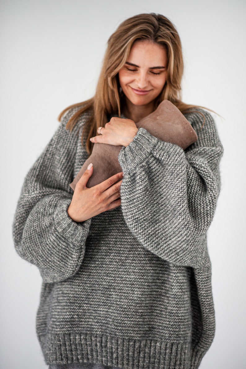 May include: A woman wearing a gray knitted sweater is holding a brown leather clutch purse.