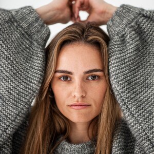 May include: A woman wearing a gray knitted sweater with a loose fit. She is looking directly at the camera with her arms raised above her head.