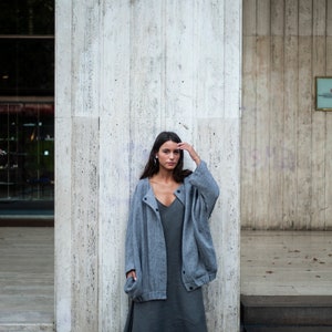 May include: A woman wearing a grey cardigan over a grey maxi dress and black heeled sandals. She is standing in front of a concrete wall.