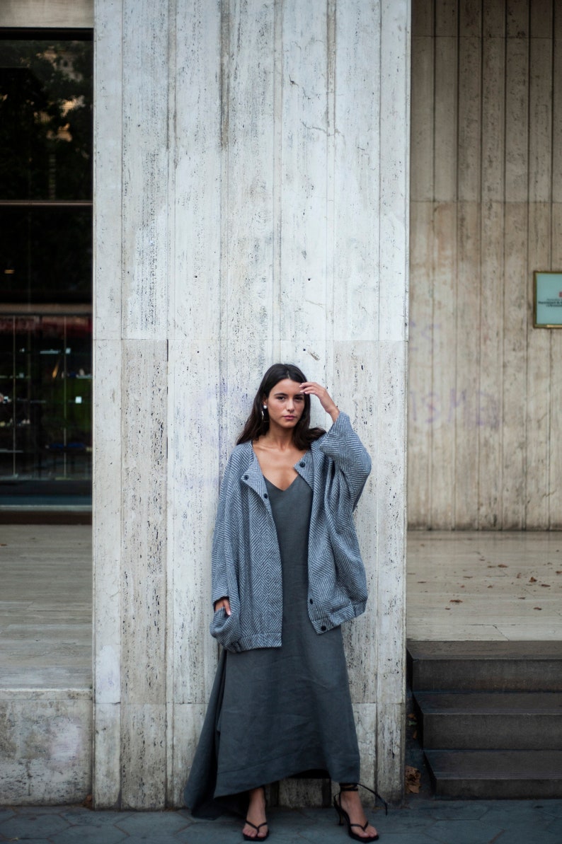 May include: A woman wearing a grey cardigan over a grey maxi dress and black heeled sandals. She is standing in front of a concrete wall.