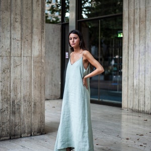 May include: A woman wearing a light blue maxi dress with thin straps and a V-neckline. She is standing in front of a glass door and a concrete wall. She is wearing black heeled sandals.