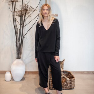 May include: A woman wearing a black long-sleeved v-neck sweater and black wide-leg pants. She is standing in front of a white wall with a wooden shelf above her head. There are dried plants in a white vase to the left of her.