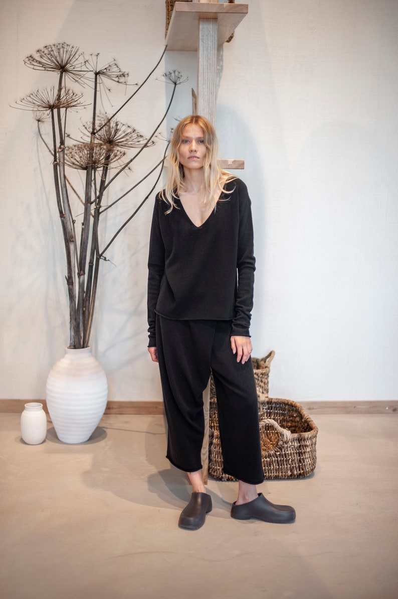 May include: A woman wearing a black long-sleeved v-neck sweater and black wide-leg pants. She is standing in front of a white wall with a wooden shelf above her head. There are dried plants in a white vase to the left of her.