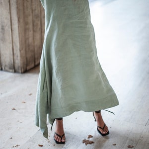 May include: A woman wearing a light green linen dress and black heeled sandals. The dress is flowing and has a relaxed fit. The sandals have thin straps and a square toe.