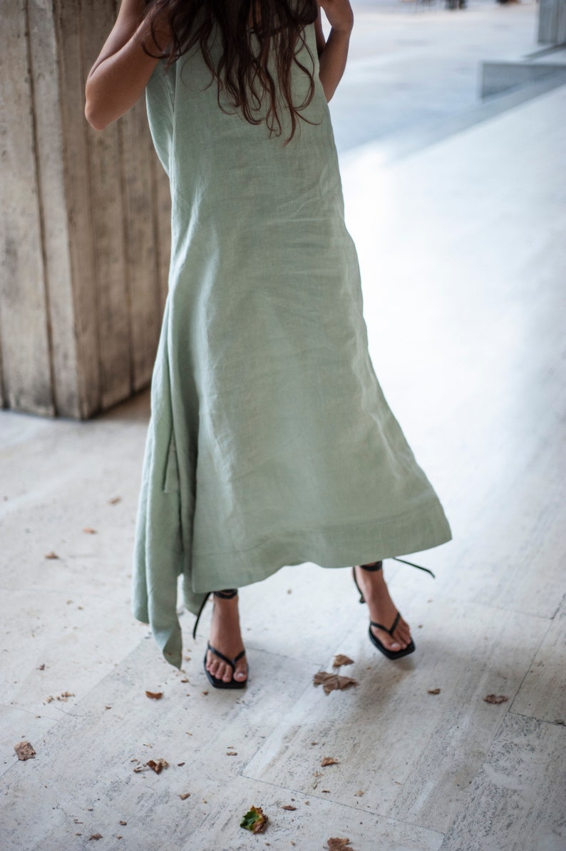 May include: A woman wearing a light green linen dress and black heeled sandals. The dress is flowing and has a relaxed fit. The sandals have thin straps and a square toe.
