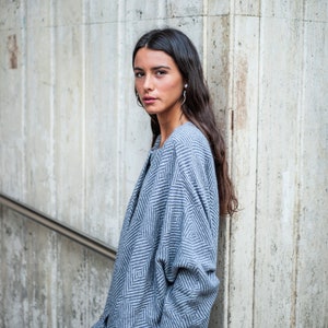 May include: A woman wearing a blue and white patterned sweater with a geometric design. She is standing in front of a concrete wall.