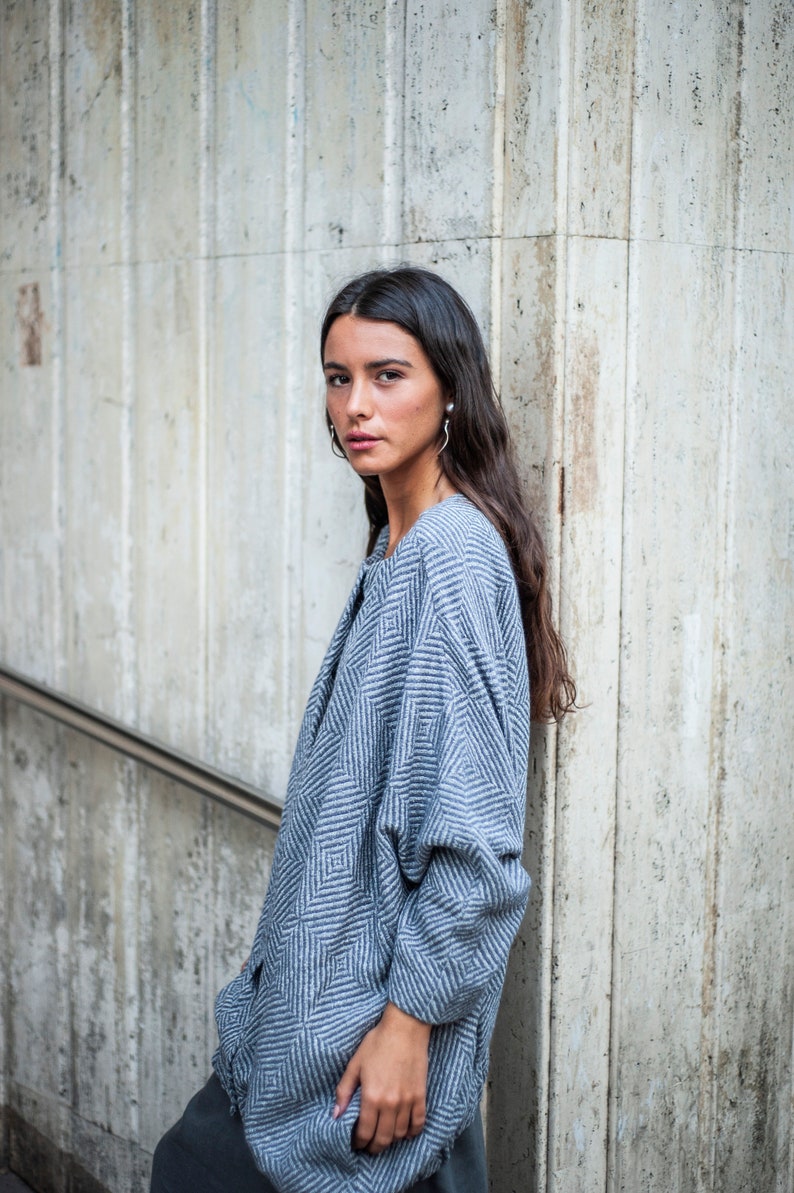 May include: A woman wearing a blue and white patterned sweater with a geometric design. She is standing in front of a concrete wall.