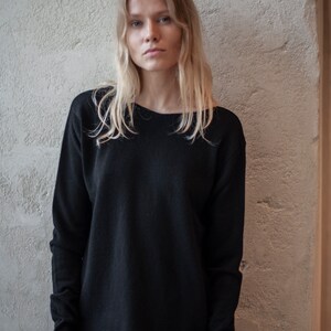 May include: A woman with long blonde hair is wearing a black long-sleeved sweater with a slightly rounded neckline. The sweater has a relaxed fit and the sleeves are slightly gathered at the wrists.