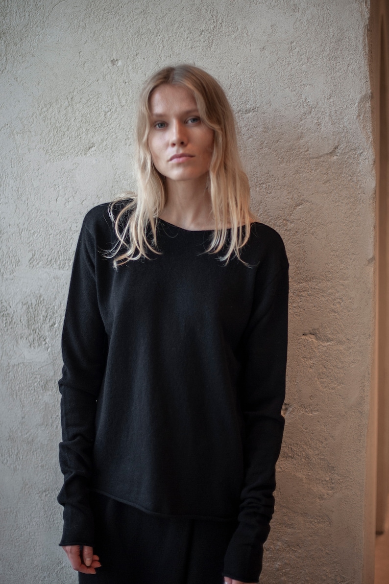 May include: A woman with long blonde hair is wearing a black long-sleeved sweater with a slightly rounded neckline. The sweater has a relaxed fit and the sleeves are slightly gathered at the wrists.