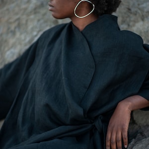 May include: A woman wearing a dark green linen wrap dress with a long sleeve and a tie belt. She is wearing large oval silver earrings.