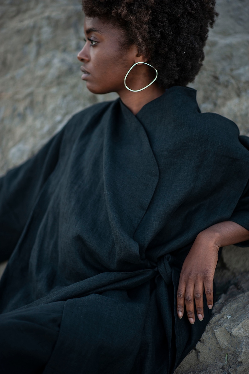 May include: A woman wearing a dark green linen wrap dress with a long sleeve and a tie belt. She is wearing large oval silver earrings.