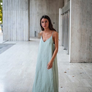 May include: A woman wearing a sage green maxi dress with thin straps and a v-neckline. She is standing in front of a concrete wall.