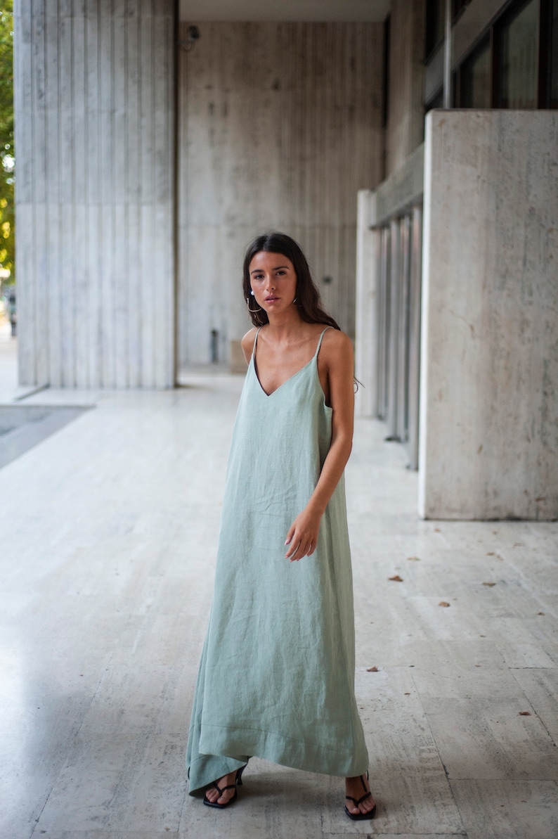 May include: A woman wearing a sage green maxi dress with thin straps and a v-neckline. She is standing in front of a concrete wall.