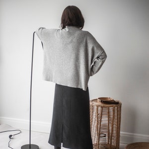 May include: A woman wearing a gray sweater and a black skirt stands in front of a white wall. She is wearing black boots and has her back to the camera. There is a wicker basket to the right of her.