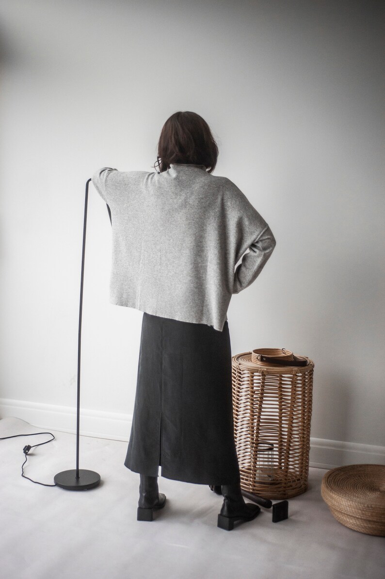 May include: A woman wearing a gray sweater and a black skirt stands in front of a white wall. She is wearing black boots and has her back to the camera. There is a wicker basket to the right of her.