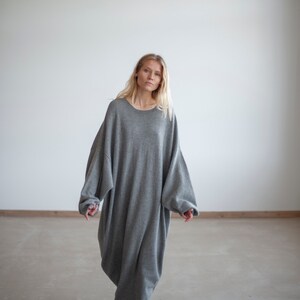 May include: A woman wearing a gray oversized sweater dress. The dress has a relaxed fit and long sleeves. She is standing on a concrete floor.