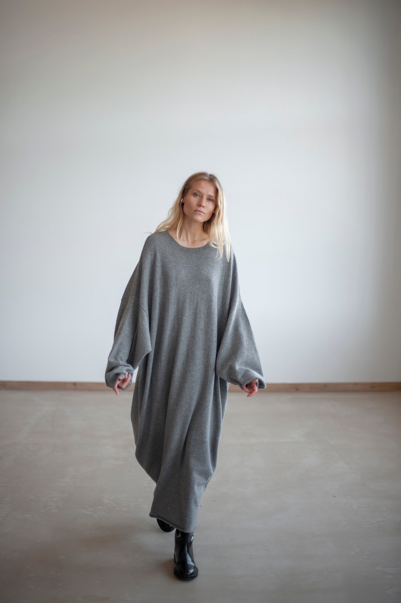 May include: A woman wearing a gray oversized sweater dress. The dress has a relaxed fit and long sleeves. She is standing on a concrete floor.