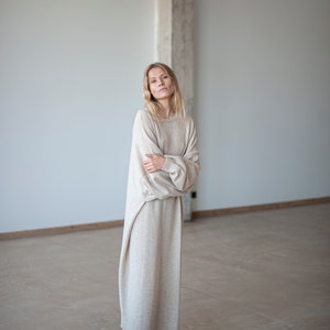 May include: A woman wearing a long, beige sweater dress with long sleeves and a relaxed fit. She is standing in a room with a white wall and a concrete pillar in the background. She is wearing black ankle boots.