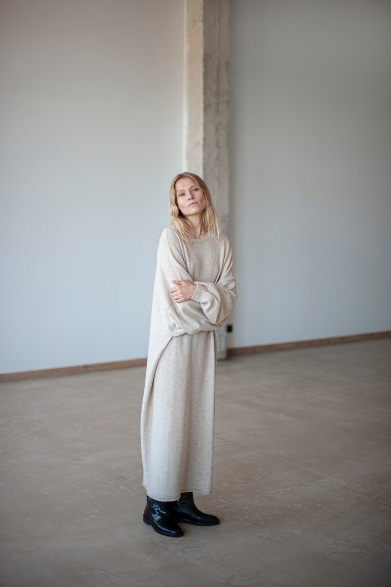 May include: A woman wearing a long, beige sweater dress with long sleeves and a relaxed fit. She is standing in a room with a white wall and a concrete pillar in the background. She is wearing black ankle boots.