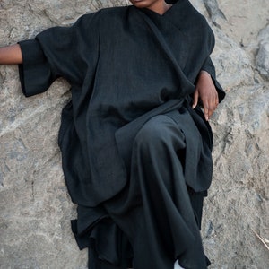 May include: A woman wearing a black linen kimono-style top and pants. She is sitting on a rock with her legs crossed and looking at the camera.