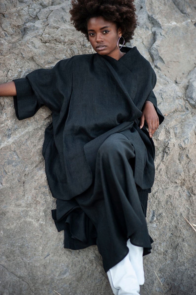 May include: A woman wearing a black linen kimono-style top and pants. She is sitting on a rock with her legs crossed and looking at the camera.