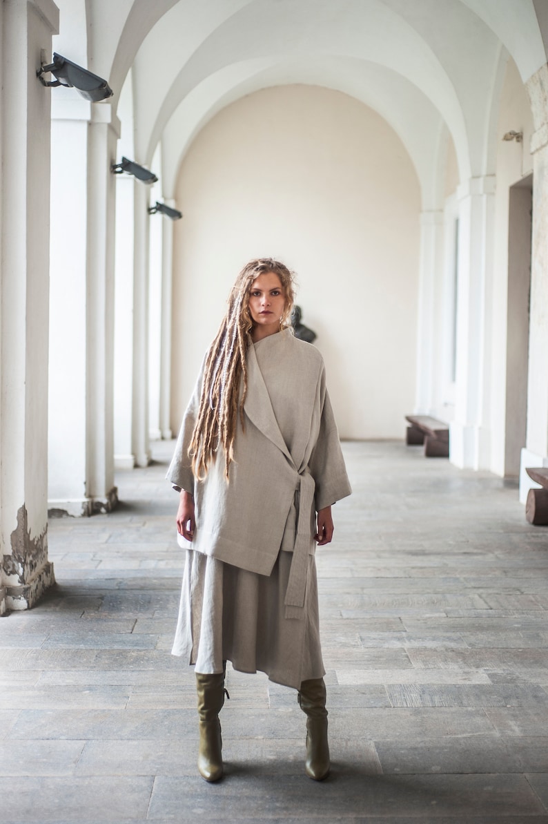May include: A woman wearing a light gray linen wrap dress and matching jacket. She has long blonde dreadlocks and is standing in a white arched hallway.