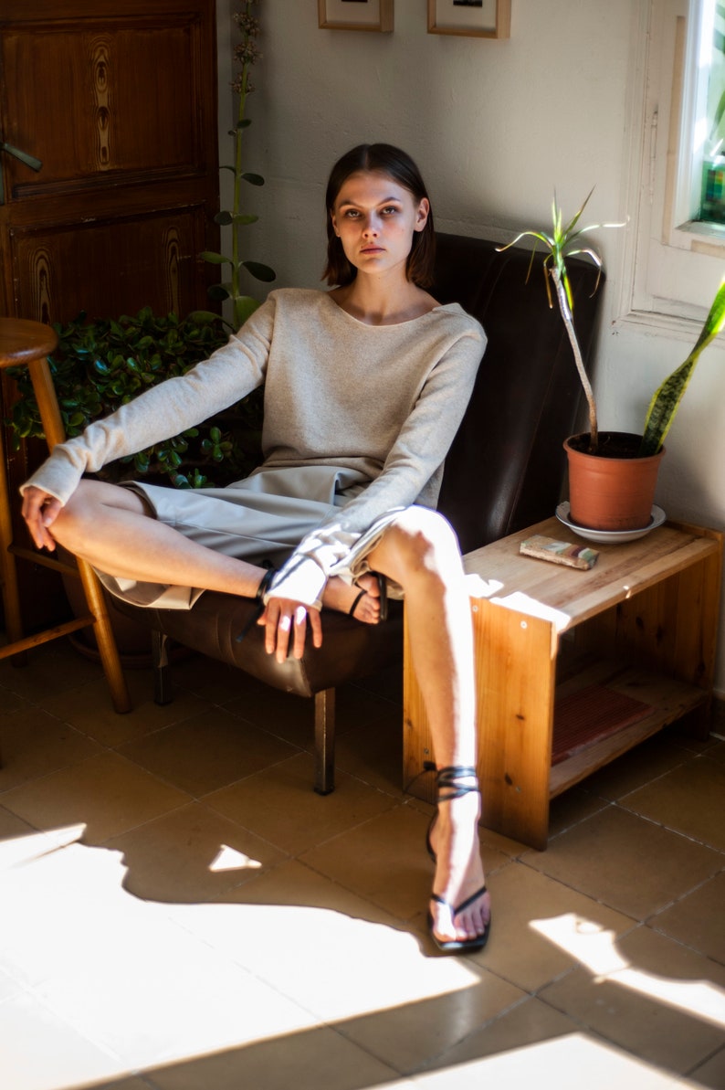 May include: A woman wearing a light beige sweater and shorts sits in a brown leather chair. She is wearing black sandals with straps. The chair is in a room with a wooden side table and plants.