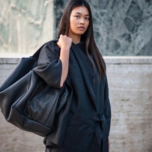 May include: A woman wearing a black long-sleeved shirt with a tie detail, carrying a large black leather tote bag.