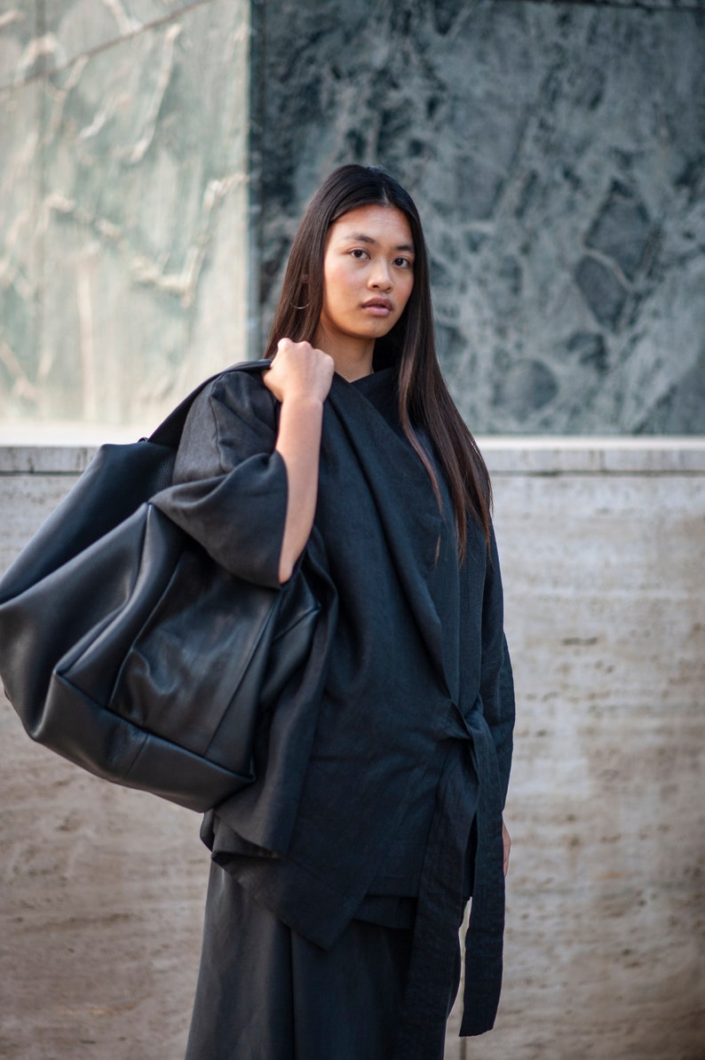 May include: A woman wearing a black long-sleeved shirt with a tie detail, carrying a large black leather tote bag.