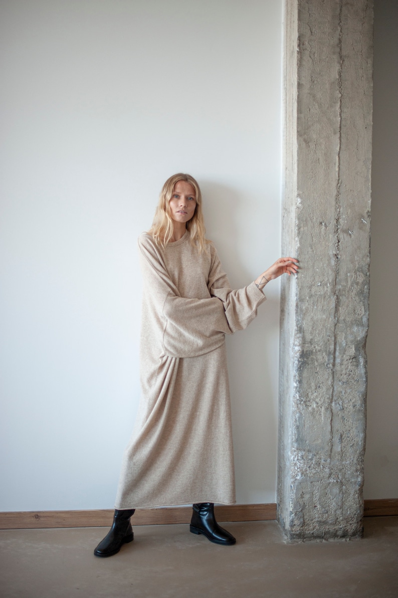 May include: A woman wearing a long, beige sweater dress with long sleeves and a relaxed fit. She is standing next to a concrete pillar and wearing black ankle boots.