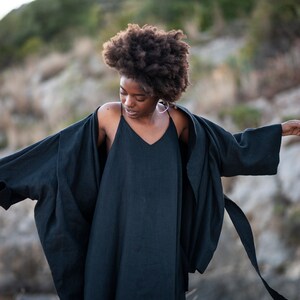 May include: A woman wearing a black linen dress and a black linen kimono with a long tie. The woman has curly dark hair and is standing in a rocky outdoor setting.