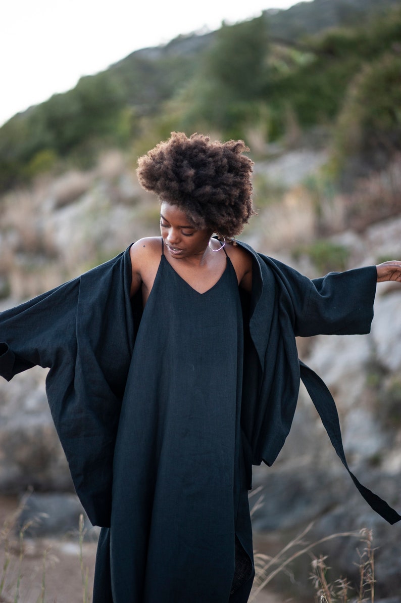 May include: A woman wearing a black linen dress and a black linen kimono with a long tie. The woman has curly dark hair and is standing in a rocky outdoor setting.