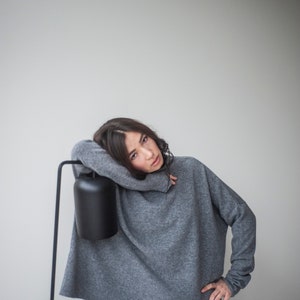 May include: A woman wearing a gray oversized sweater with a black lampshade behind her.