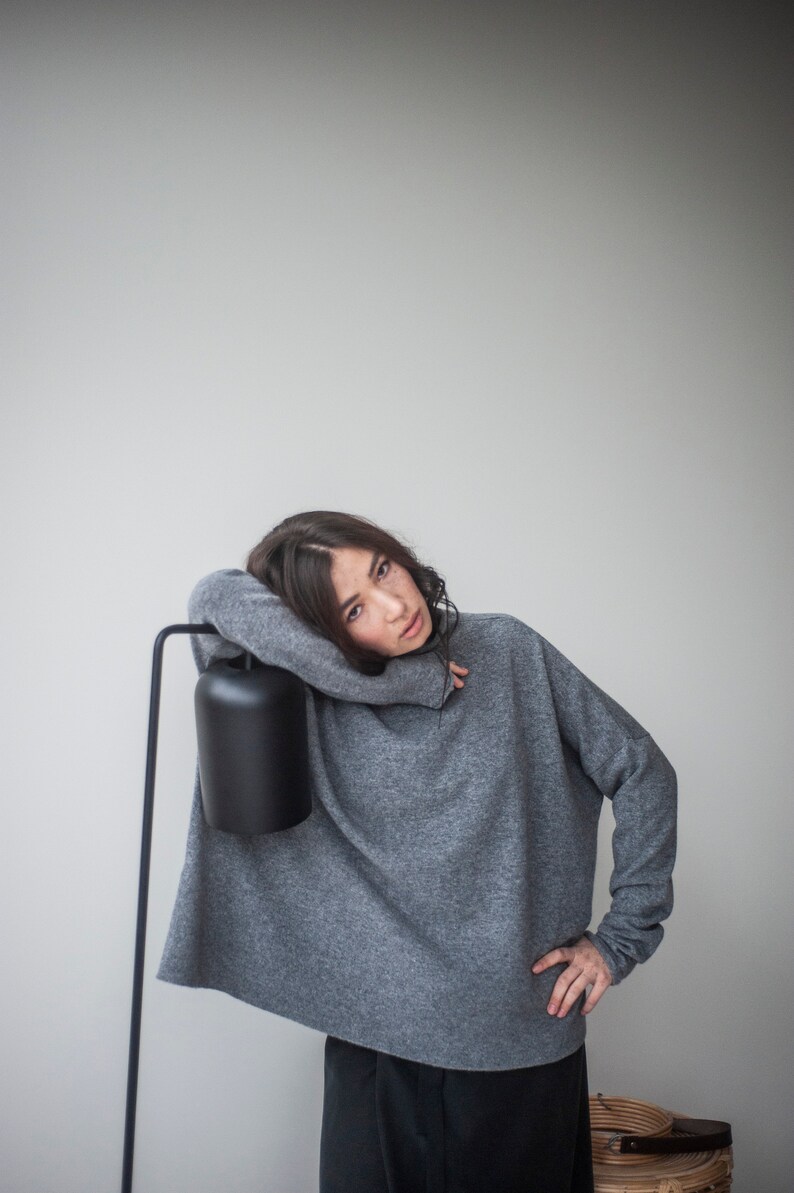 May include: A woman wearing a gray oversized sweater with a black lampshade behind her.