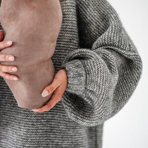May include: A person is holding a large, brown, ceramic vase with a textured surface. The person is wearing a gray knitted sweater with a loose fit.