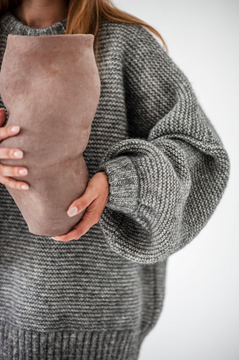 May include: A person is holding a large, brown, ceramic vase with a textured surface. The person is wearing a gray knitted sweater with a loose fit.