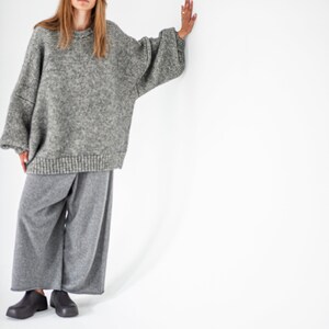 May include: A woman wearing a gray oversized sweater and gray wide-leg pants. She is standing with her right hand on a white wall.