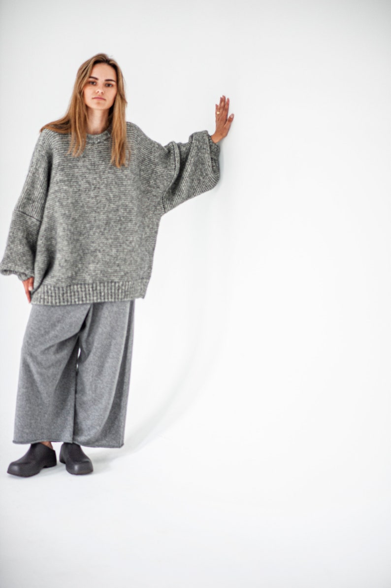 May include: A woman wearing a gray oversized sweater and gray wide-leg pants. She is standing with her right hand on a white wall.